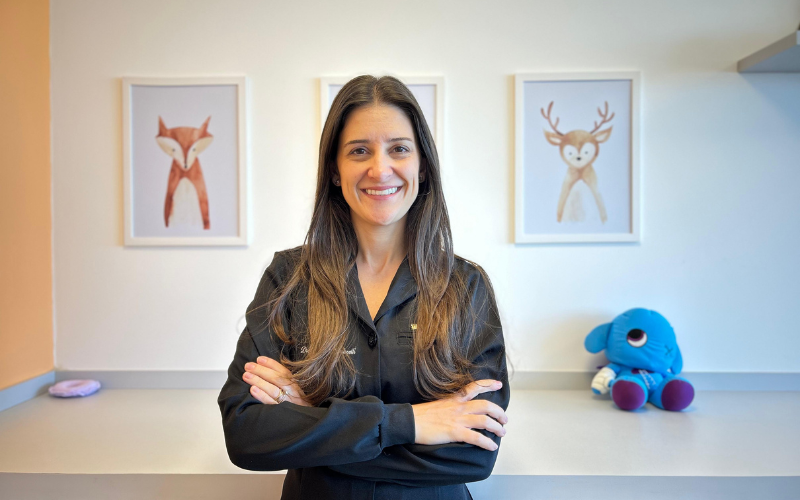 Mulher,medica sorridente de cabelos longos, vestindo camisa preta, em ambiente decorado com tema infantil, com quadros de animais fofos e um brinquedo de pelúcia azul ao fundo.