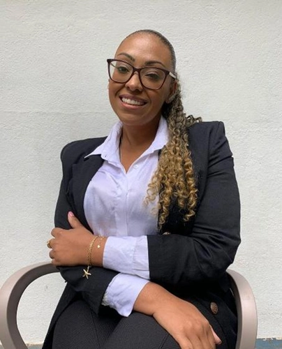 Mulher negra de terno preto e camisa branca e cabelos cacheados loiros.