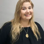 Mulher branca e loira com blusa de manga preta em um fundo cinza e sorrindo com a cabeça levemente inclinada