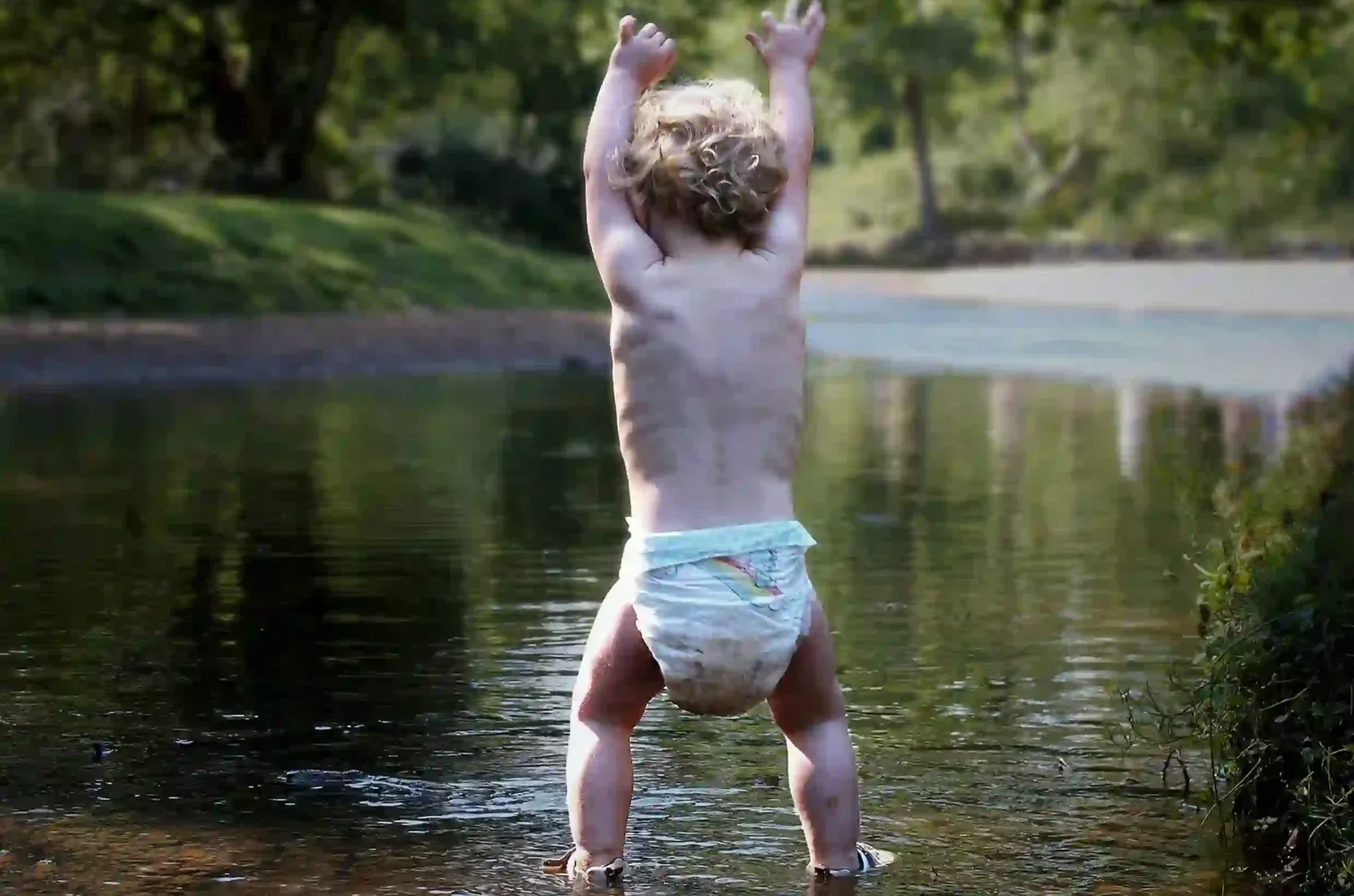 Criança apenas de fralda, com os pés no rio, cercada por árvores.