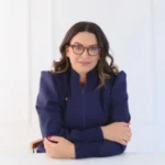 A imagem mostra uma mulher com cabelos castanhos ondulados, usando óculos de grau e um blazer azul escuro com mangas bufantes. Ela está sentada à mesa, com os braços cruzados e um leve sorriso no rosto. O fundo é branco e minimalista.