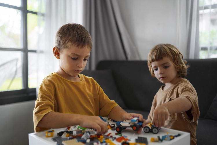 Dois meninos pequenos brincam com peças de montar sobre uma mesa branca. Estão sentados em um sofá e parecem concentrados. Ambos usam camisetas em tons de marrom claro.