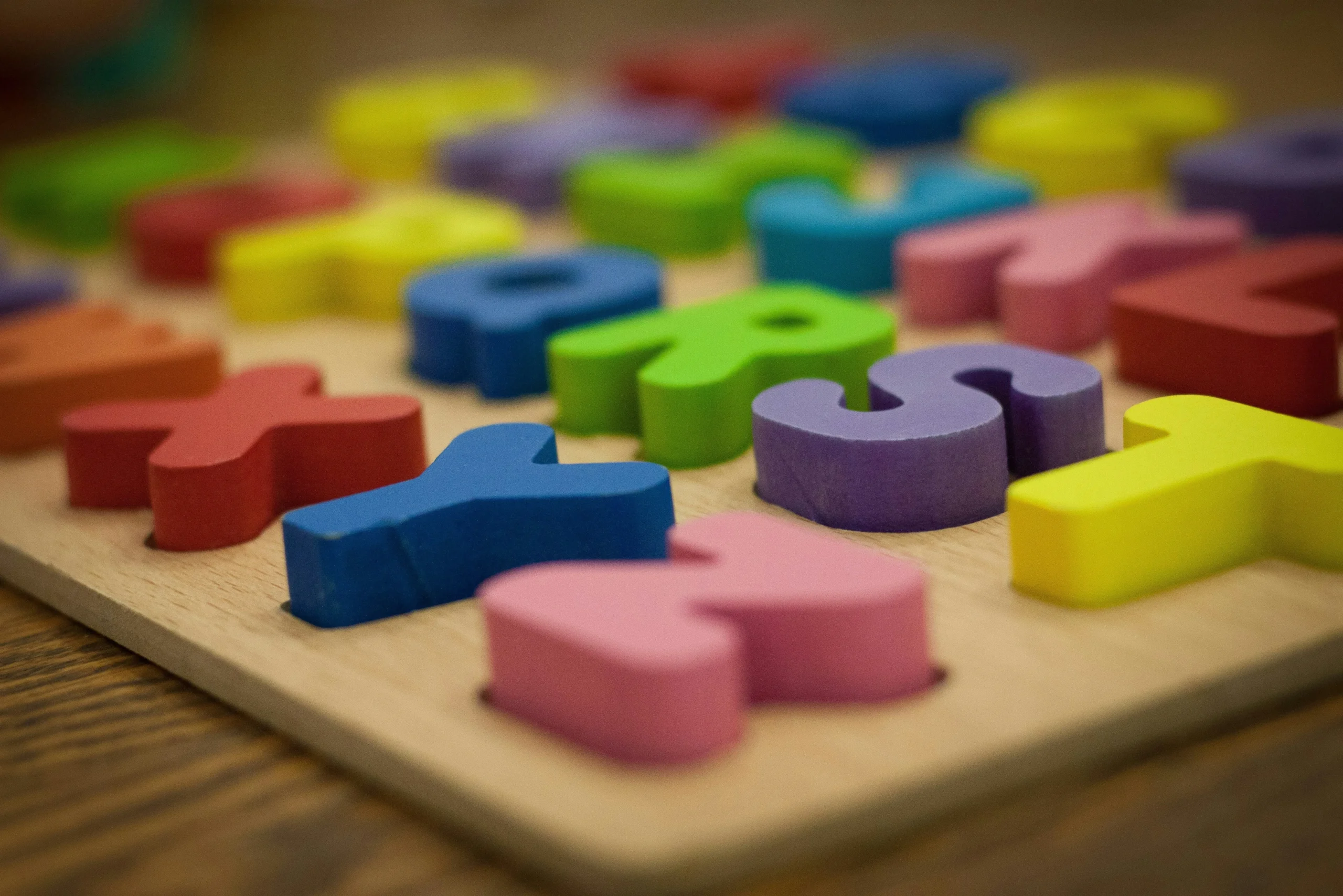 Jogo de tabuleiro tipo Quebra-cabeça de madeira com peças de letras do alfabeto em cores vibrantes, como vermelho, azul, verde, amarelo, rosa e roxo, dispostas sobre uma superfície de madeira.