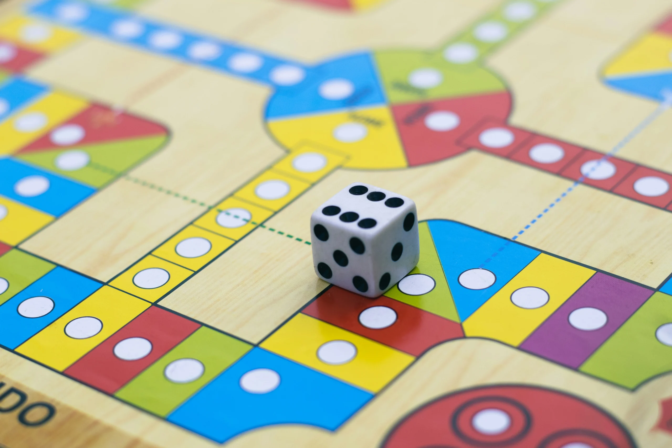 Jogo de tabuleiro colorido com quadrados e bolas brancas. Muita luz natural e um dado pousa o jogo com o número 6 para cima.