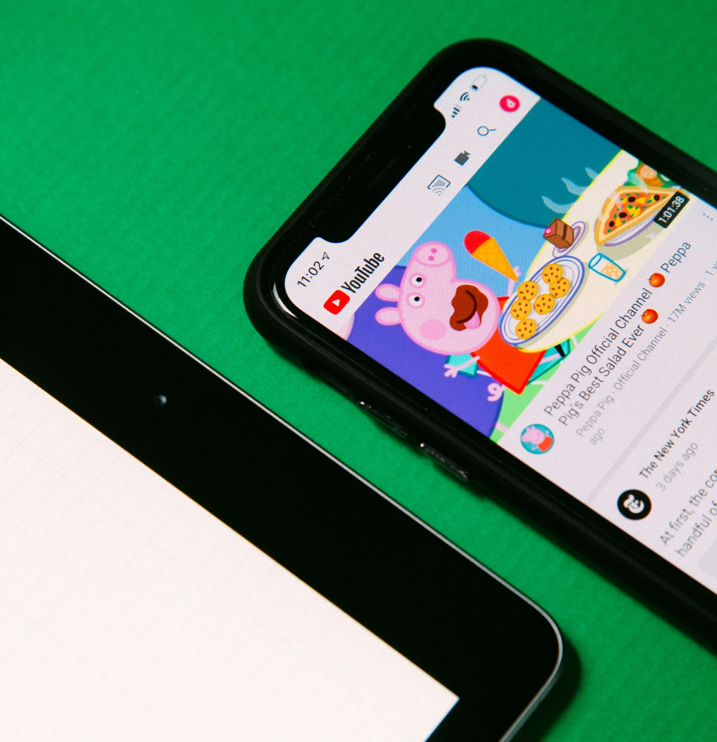 Mesa verde com um tablet e um iphone ligado no Youtube com um vídeo do desenho animado "Peppa Pig" onde a personagem principal está sentada à mesa com a boca bem aberta a comer uma sopa e massa.