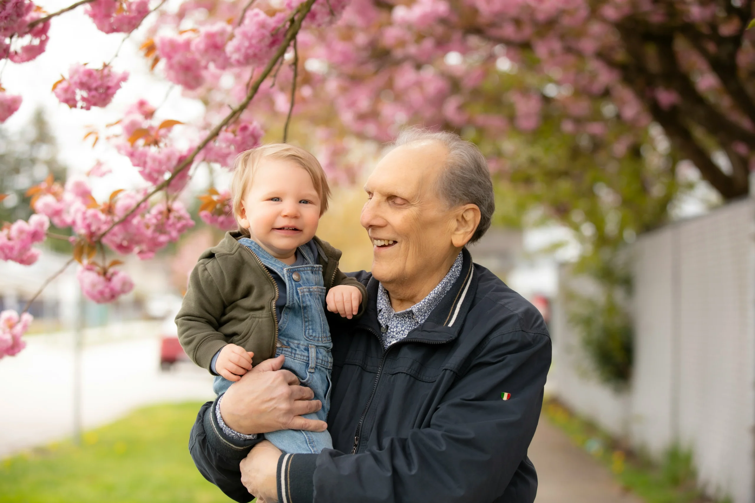 Homem idoso segura uma criança pequena nos braços enquanto ambos sorriem sob flores de cerejeira cor-de-rosa em um dia claro. A interação mostra conexão entre gerações. Acessibilidade: espaço ao ar livre plano e arborizado, ideal para convivência familiar com segurança para todas as idades.