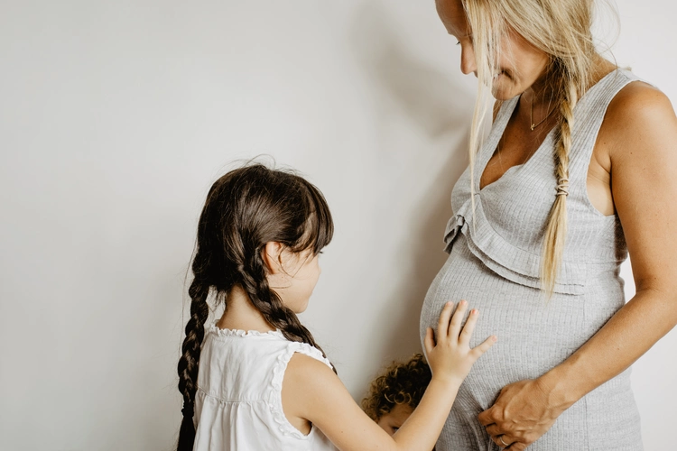 Uma menina, com delicadeza, coloca a mão na barriga da sua mãe, que está grávida. As duas têm tranças no cabelo e vestem roupas de cores claras.
