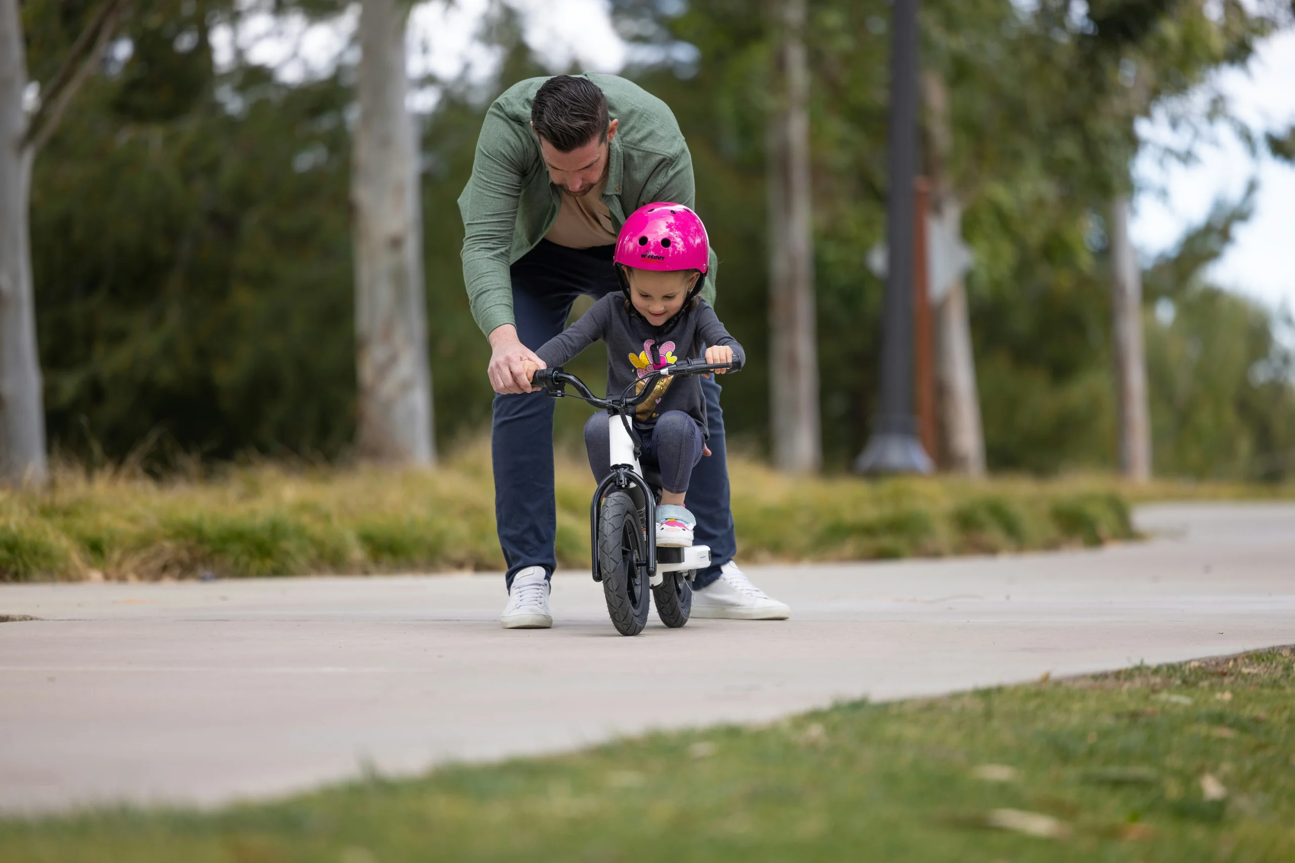Menina de 1 a 2 anos de idade andando de bicicleta de equilibrio roxa clara na rua com um capacete rosa choque. Bicicleta sem pedais de equilíbrio. Está sorridente numa rua citadina com o pai.