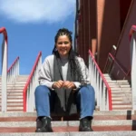 Mulher sentada no meio de uma escada externa, com corrimãos vermelhos. Ela tem cabelos longos com tranças, usa suéter cinza, cachecol escuro, calça jeans e botas pretas. Está sorrindo, com as mãos apoiadas nos joelhos. Ao fundo, céu azul e parte de um prédio de tijolos aparentes.