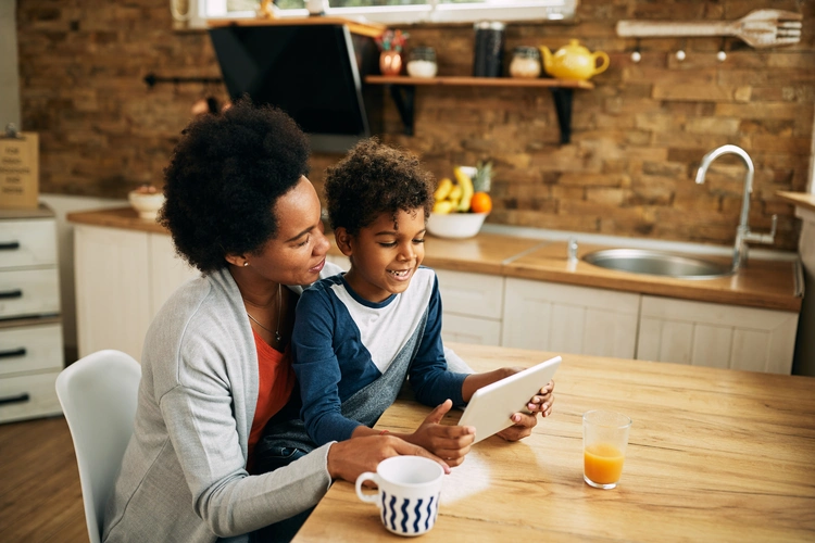 Mãe negra e filho sorrindo enquanto usam um tablet juntos na cozinha, com suco e frutas sobre a mesa.