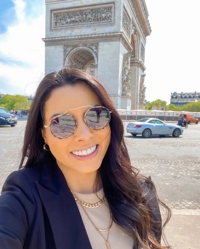 Mulher de pele morena, cabelo longo, liso e castanho escuro, sorrindo em frente ao Arco do Triunfo, em Paris, usando óculos de sol, blazer preto e colar dourado.