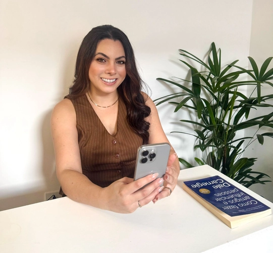 Mulher jovem de cabelos longos e castanhos, sorrindo, segurando um celular em um ambiente claro e minimalista. Sobre a mesa há um livro intitulado 'Como Fazer Amigos e Influenciar Pessoas' e uma planta decorativa ao fundo.