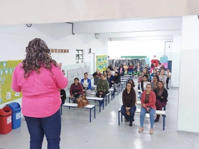 Mulher negra de cabelos cacheados castanhos, usando camisa rosa, vista de costas enquanto fala para um grupo de cerca de 20 pessoas sentadas em bancos dentro de uma sala clara e ampla. O público acompanha atentamente a palestra, com murais coloridos e lixeiras ao fundo.