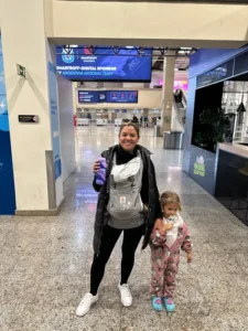 mulher com um bebê menor de um ano no canguru e segurando a mão de uma menina menor em um aeroporto