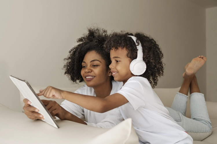 Mãe e filho negros sorridentes interagem juntas em um tablet, enquanto a criança usa fones de ouvido brancos. Cena de vínculo familiar e uso consciente da tecnologia em casa.