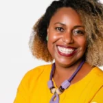 Mulher negra sorridente com cabelo cacheado com mechas loiras nas pontas. Ela veste uma blusa amarela vibrante e um colar roxo com detalhes grandes. Está posando diante de um fundo claro.