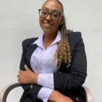 Mulher negra sentada em uma cadeira, sorrindo, vestindo blazer preto e camisa social branca, com cabelo preso e óculos de grau, em frente a uma parede clara.