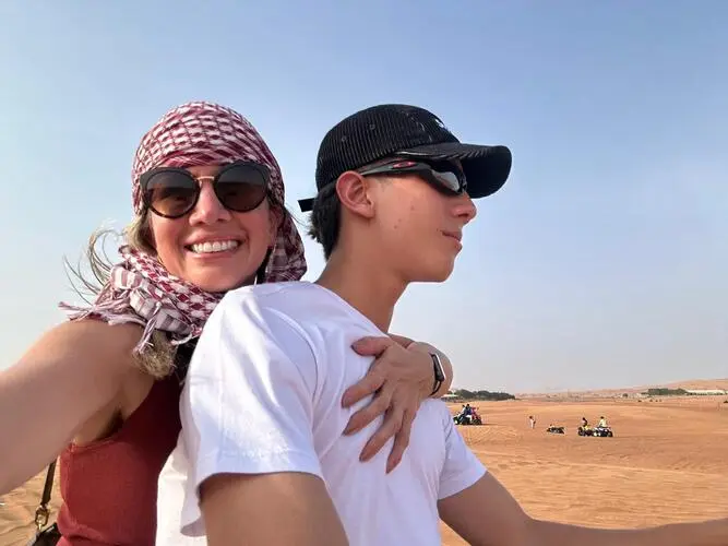 Uma mulher usando um lenço na cabeça, abraçando com apenas uma mão um adolescente que usa boné, ambos brancos e usando óculos escuros, a imagem tem teor maternal em uma paisagem arenosa.