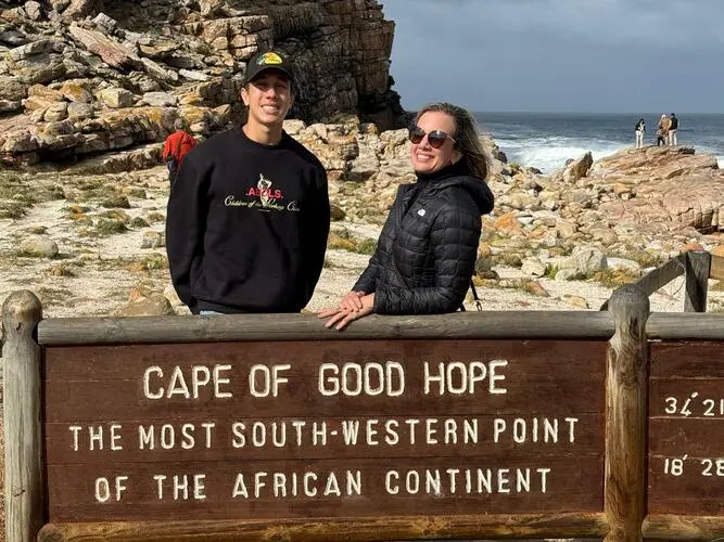 homem adolescente e mulher branca, vestidos com roupas pretas, aparecem atrás de uma placa em meio a uma praia com rochas e pedras