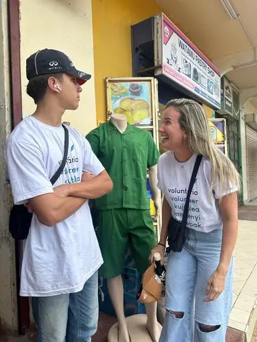 Mulher loira aparentando 40 anos, rindo a ponto de inclinar o corpo para um adolescente de boné e braços cruzados.