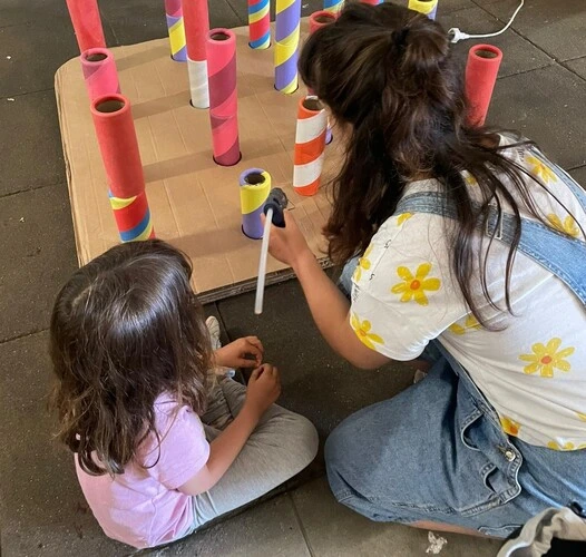 mulher branca e filha pequena aparecem de costas, sentadas no chão, enquanto trabalham com elementos criativos. A mulher veste blusa branca com flores amarelas e uma jardineira jeans, e a criança pequena camiseta rosa e calça cinza
