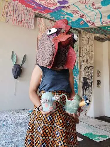 mulher de blusa azul e vestindo uma cabeça de galinha aparece segurando duas esculturas