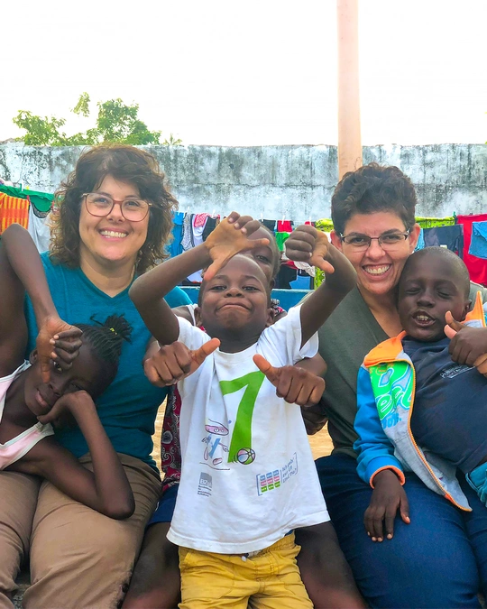 duas mulheres brancas de blusa azul e cinza e óculos aparecem sorrindo com crianças negras
