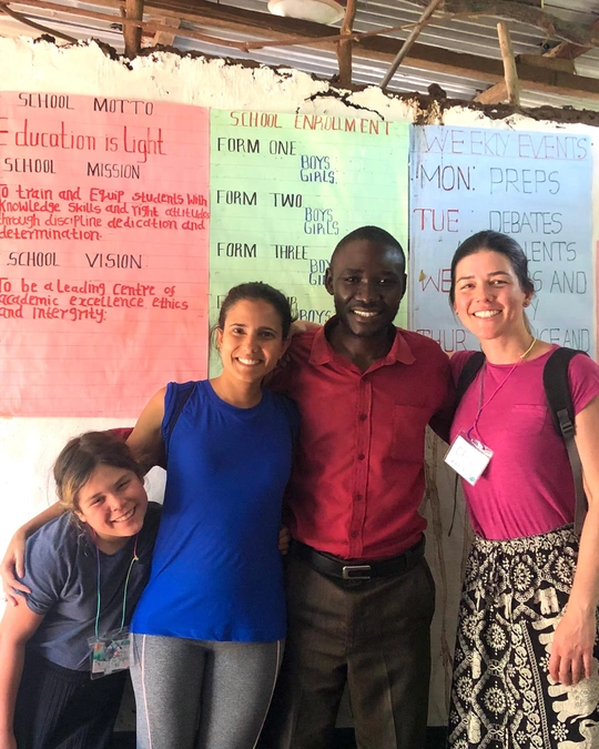 três pessoas brancas, duas mulheres e uma criança, abraçam homem negro. Todos estão sorrindo. Eles aparecem em frente a um mural com cartazes e descrições