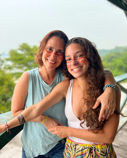 mulher de regata azul e menina adolescente de regata branca, ambas de pele branca, aparecem sorrindo em ponte.