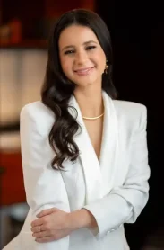 "Mulher jovem com cabelo castanho-escuro e longo, penteado em ondas suaves. Ela está sorrindo levemente e usa um blazer branco elegante, com uma corrente dourada no pescoço e brincos dourados. Está em um ambiente interno com iluminação suave e fundo desfocado. Ensina como escovar dentes, é dentista."