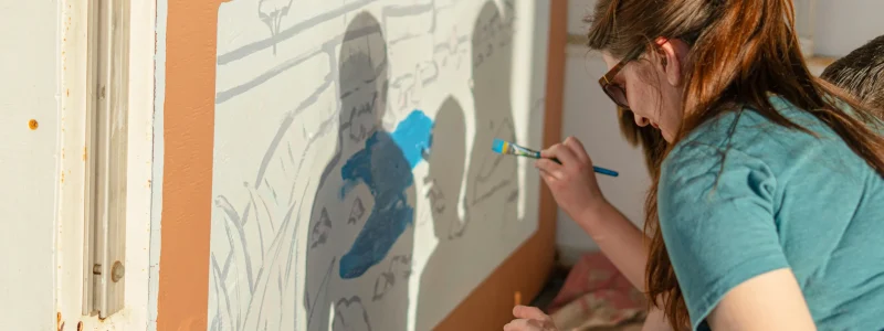 Mãe pinta obra de arte na parede com a filha, realçando a conexão entre pais e filhos. A sombra das pessoas é refletida na parede cor de salmão. Há luz natural e a mãe, de cabelos compridos castanhos, veste uma camisa azul esverdeada.