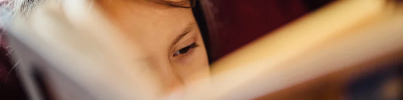 menina no fundo de cabelo castanho escuro lendo compenetrada. Livro no plano da frente desfocado ocupando quase a imagem total. Há um janela no fundo à refletir luz solar e com uma cortina vermelha escura ao lado.