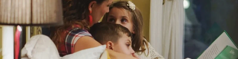 Numa casa com parede amarela, grinalda na parede e janela com cortina branca apanhada, encontra-se uma mãe com dois filhos sentada a ler um livro para eles numa cama com muitas almofadas brancas e com uma mesinha com um abajur ao lado cor castanho. A mulher tem cabelos compridos loiros apanhados e as crianças de pijama azul.