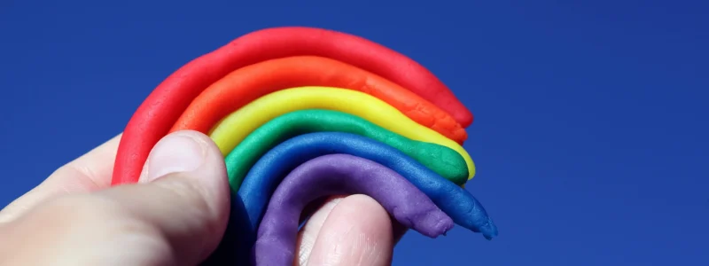 Uma mão segura delicadamente um arco-íris feito de massinha de modelar, com cores vibrantes em um fundo azul intenso do céu. As tiras coloridas estão dispostas em camadas, curvando-se para formar o arco característico.