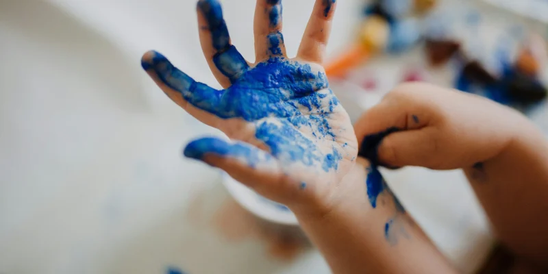 Mão de uma criança coberta de tinta azul, com fundo desfocado mostrando outros materiais de pintura.
