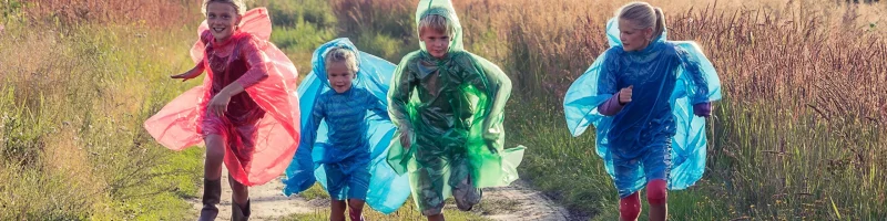 Quatro crianças estão correndo por uma estrada de terra no campo, vestindo capas de chuva coloridas (vermelha, azul e verde). Elas parecem felizes e brincando, enquanto a luz do sol ilumina a paisagem ao redor. Ao fundo, há uma casa branca com um telhado escuro e vegetação alta.